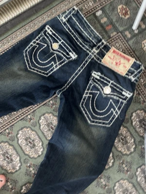 True Religion mörkblå jeans - Säljer ett par mörkblå jeans från True Religion med kraftiga vita kontrastsömmar och ikoniska bakfickor med stora broderade detaljer. Jeansen har silverfärgade knappar och patch på bakfickan. Materialet är denim och passformen är klassisk.
