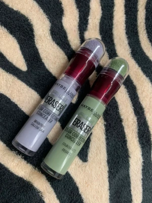 Maybelline Eraser Color Correcting Concealer - Två Maybelline Eraser Color Correcting Concealers, en i lila och en i grön nyans. Båda har en praktisk applikator med svamp och genomskinlig plasttub med vinröd kork. Perfekt för att neutralisera olika hudtoner och korrigera ojämnheter.