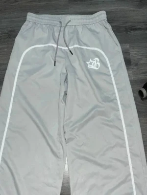 Gray Beaudrm Y2k Graphic Baggy Pants - Snygga grå mjukisbyxor från Billionaire Boys Club med vita detaljer längs benen och logga på ena låret. Byxorna har resår i midjan och dragsko för skön passform. Perfekta för en chill och avslappnad stil.