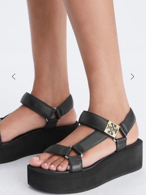 Svarta platåsandaler Tory Burch - Snygga svarta sandaler med platåsula från Tory Burch. Remmar i läder med guldfärgad logodetalj på sidan. Justerbara spännen och modern, chunky design som ger extra höjd. Perfekta för dig som gillar trendiga och bekväma skor.