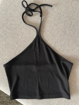 Svart ribbad halterneck linnetopp - Snygg svart ribbad linnetopp med halterneck och knyt i nacken. Toppen är croppad och har en stilren, minimalistisk look som passar perfekt till jeans eller kjol. Materialet är mjukt och stretchigt för en bekväm passform.
