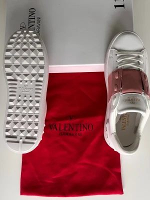 Valentino Garavani vita sneakers rosa detalj - Snygga vita sneakers från Valentino Garavani med breda rosa läderdetaljer över snörningen. Skorna har klassisk låg profil, vit sula med mönster och guldfärgad logga på plösen. Tillverkade i läder för en lyxig känsla.