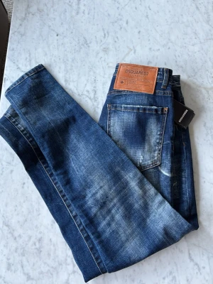 Blå distressed jeans från Dsquared2 - Säljer ett par blå jeans från Dsquared2 med slitna detaljer och tvättad look. Jeansen har raka ben, klassisk femficksmodell och läderpatch bak i midjan. Materialet är denim i bomull och de har en cool, trendig vibe.