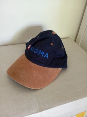 Ny SIGMA keps - Säljer en snygg keps från SIGMA med mörkblått tyg och broderad logga framtill. Skärmen är i ljusbrunt mockaliknande material och kepsen har klassisk sexpanelsdesign med metallöljetter. Perfekt för dig som vill ha en stilren accessoar.