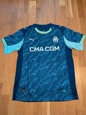 Helt ny Blå Marseille fotbollströja Puma. Storlek. S.M.L - Säljer en blå fotbollströja från Puma för Olympique de Marseille. Tröjan har digitalt mönster i olika blå nyanser, korta ärmar med ljusblå och gula detaljer samt klubbmärke och Puma-logga på bröstet. Perfekt för fotbollsträning eller match!