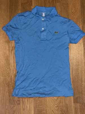 Blå pikétröja från Lacoste - Klassisk blå pikétröja från Lacoste med korta ärmar, krage och knappar framtill. Tröjan har den ikoniska krokodilloggan på bröstet och är tillverkad i ett mjukt bomullsmaterial. Perfekt för en avslappnad och sportig stil.