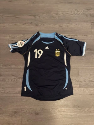 Argentina Messi fotbollströja #19 - Mörkblå Argentina fotbollströja från Adidas med ljusblå och vita detaljer. Nummer 19 och Messi på ryggen, samt landslagets emblem på bröstet. Korta ärmar och sportig passform, perfekt för fotboll eller träning.