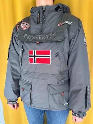 Anorak fodrad - Fodrad anorak från Geographical Norway med stor norsk flagga framtill, broderade patches och dragkedjor. Jackan har huva, flera fickor och justerbara muddar. Sitter som en S/M💫