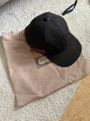 Svart gucci keps - Snygg svart keps Gucci från med diskret gg logomönster över hela kepsen. Klassisk böjd skärm och ikoniska gröna och röda ränder på sidan. Kepsen är tillverkad i tyg och har en stilren look som passar perfekt till streetwear.