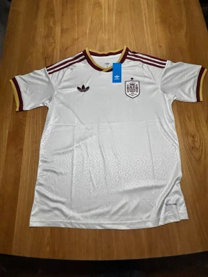 Vit Spanien fotbollströja Adidas - Vit Spanien fotbollströja från Adidas med vinröda detaljer på krage, ärmslut och axlar. Tröjan har korta ärmar, broderad logga och klubbmärke på bröstet samt subtilt mönster i tyget. Perfekt för fotboll och träning. Längd 72cm, bredd 51cm