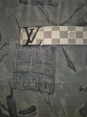  rutigt bälte i beige - Snyggt bälte  med klassiskt rutigt mönster i beige och grått. Bältet har en stor -logga i metall som spänne och är tillverkat i skinn. Perfekt accessoar för att lyfta din outfit.