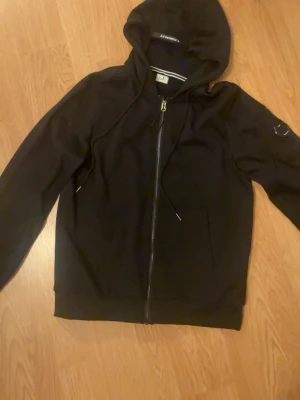 Svart hoodie  - Svart hoodie med dragkedja. Klassisk modell med huva, fickor framtill och ikonisk linsdetalj på ärmen. Mjuk bomullskänsla och stilren design, perfekt för en avslappnad streetwear-look.