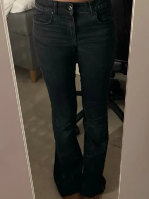 Svarta bootcut jeans med låg midja - Säljer ett par svarta bootcut jeans med låg midja och klassiska fem fickor. Jeansen har en snygg utsvängd passform nertill och är perfekta för dig som gillar en trendig siluett. Materialet är stretchigt denim som sitter bekvämt hela dagen. Dock har de ett litet hål där nere.