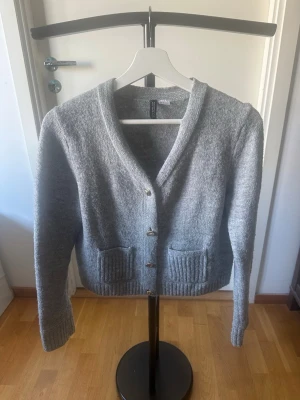 Grå kofta från H&M med fickor - Mysig grå kofta från H&M med v-ringning och knappar framtill. Koftan har två framfickor och ribbade muddar vid ärmslut och nederkant. Perfekt för lager-på-lager och enkel att matcha med olika outfits.Nopprig på vissa ställen