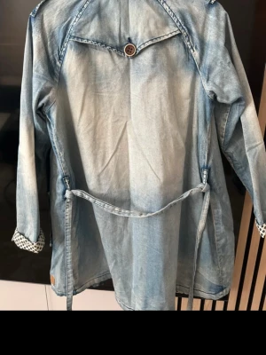 Nümph, Ljusblå jeansjacka  - Snygg ljusblå jeansjacka från Nümph med knytband i midjan och dekorativ knappdetalj på ryggen. Jackan har långa ärmar och ett unikt mönster på insidan av ärmsluten. Perfekt för dig som vill ha en avslappnad men trendig look.
