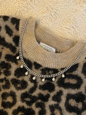 Silverhalsband med pärldetaljer - Stilrent halsband i silverfärgad rostfritt med sju små hängande pärlor. Kedjan är platt och bred, vilket ger ett modernt intryck. Perfekt accessoar för att lyfta din outfit med en touch av elegans.