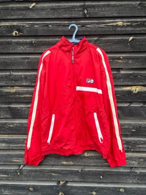 Röd vindjacka från FILA med vita detaljer - Riktigt snygg röd vindjacka från FILA med vita ränder längs ärmarna och vita dragkedjor. Jackan har hög krage, två fickor framtill och en liten FILA-logga på bröstet. Perfekt för en sportig och retro look.