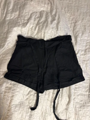 Svarta linne shorts med knytskärp - Svarta shorts med knytskärp i midjan. Modellen har uppvikta benslut och sidofickor. Perfekta för en avslappnad och trendig look. Materialet ser ut att vara mjukt och bekvämt, troligen bomull eller viskos.