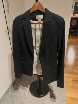 Svart kavaj från H&M - Stilren svart kavaj från H&M med klassisk passform och smala slag. Kavajen har två fickor framtill och en bröstficka. Insidan är fodrad med ett vitt tyg med tunna ränder. Perfekt för en snygg och uppklädd look.  Se bild med H&M loggan där det är en missfärgning, i övrigt gott skick.