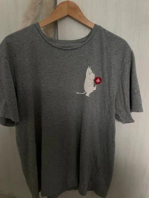 Moomin X Wood Wood T-Shirt - Grå t-shirt med ett vitt Mumin-tryck på bröstet där Mumin sparkar en röd boll med ett vitt A-märke. T-shirten har rund hals och korta ärmar, perfekt för en avslappnad stil.