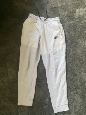 Vita Nike mjukisbyxor med dragsko - Snygga vita mjukisbyxor från Nike med svart logga på låret och elastisk midja med dragsko. Byxorna har fickor på sidorna och muddar vid bensluten. Perfekta för en avslappnad och sportig stil.