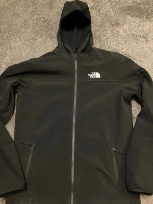 Svart vindjacka från The North Face - Snygg svart vindjacka från The North Face med huva och hel dragkedja framtill. Jackan har två sidofickor med dragkedja och logga på bröstet samt ryggen. Tillverkad i ett lätt och vindtätt material som passar perfekt för blåsiga dagar.