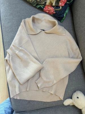 Beige stickad tröja med krage - Säljer en beige stickad tröja med bred krage och långa ärmar. Tröjan har ribbstickade muddar och nederkant, samt en avslappnad passform. Perfekt för lager-på-lager och enkel att matcha med jeans eller kjol.