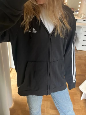 Svart Adidas hoodie med dragkedja - Svart hoodie från Adidas med dragkedja framtill, vita klassiska ränder längs ärmarna och Adidas-logga på bröstet. Stor ficka framtill och huva med snören. Perfekt till jeans eller joggers för en chill look.