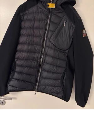 Svart pufferjacka från Parajumpers - Snygg svart pufferjacka från Parajumpers med huva, dragkedja och flera fickor. Jackan har quiltad framsida och rygg, samt släta ärmar. Märkets logga på ärmen och gul detalj vid halsen. Dragkedjan på bröstet är trasigt
