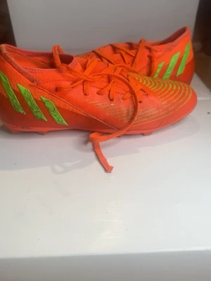 Adidas orange fotbollsskor - Säljer ett par grymma fotbollsskor från Adidas i en riktigt snygg orange färg med neongröna detaljer och klassiska ränder på sidan. Skorna har snörning och är tillverkade i syntetmaterial som ger bra grepp och känsla på planen. Perfekta för dig som vill sticka ut på matchen!