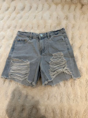 Ljusblåa denimshorts med slitningar - Snygga ljusblåa jeansshorts med rejält slitna detaljer framtill och råa kanter. Klassisk femficksmodell med bälteshällor och knappstängning. Perfekta för sommaren och ger en cool, avslappnad vibe.