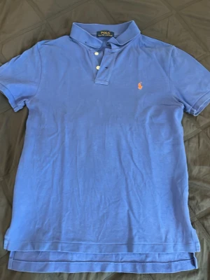 Blå pikétröja från Polo Ralph Lauren - Klassisk blå pikétröja från Polo Ralph Lauren med vit knappslå och den ikoniska broderade loggan i orange på bröstet. Tröjan har korta ärmar, krage och är tillverkad i bomull för en bekväm känsla. Perfekt till jeans eller shorts. Storlek 14-16