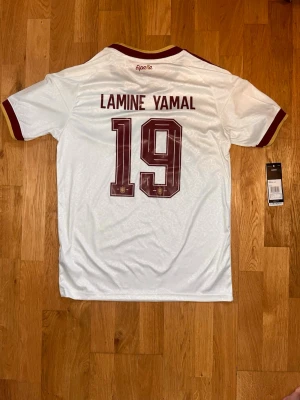 Spanien Lamine Yamal matchtröja - Vit Spanien matchtröja från Adidas med vinröda detaljer, nummer 19 och Lamine Yamal på ryggen. Tröjan har officiella tryck, broderad landslagssköld och Adidas-logga på bröstet samt patchar på ärmarna. Tillverkad i lätt och ventilerande material. Använt en gång 10/10 skick, köpt i Spanien men jag gillar inte den. Priest går att diskuteras!!!