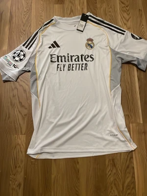 Real Madrid matchtröja Adidas vit - Säljer en vit Real Madrid matchtröja från Adidas med svarta detaljer och klubbmärke på bröstet. Tröjan har korta ärmar, svarta ränder på axlarna och sponsortexten 'Emirates Fly Better' på framsidan. Tillverkad i lätt och ventilerande material.