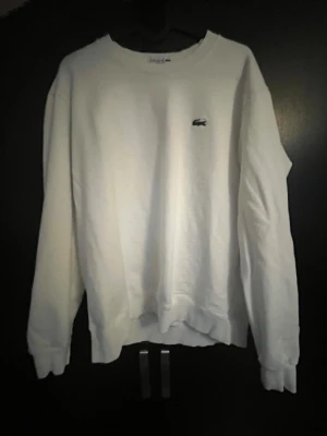 Vit sweatshirt från Lacoste - Vit sweatshirt från Lacoste med klassisk rund halsringning och broderad krokodil-logga på bröstet. Tröjan har ribbade muddar vid ärmslut och nederkant. Perfekt för en clean och stilren look. Den är storlek M men passar jätte bra för S också! Den har tagg och påse med!