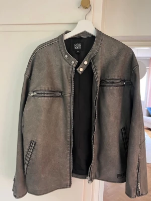 Grå skinnjacka från BDG - Snygg grå skinnjacka från BDG outfitters med raka linjer och coola dragkedjedetaljer på bröstet och vid fickorna. Jackan har en stilren krage med tryckknappar och är perfekt för dig som vill ha en edgy look. Passar till många olika outfits och har en modern, avslappnad passform.