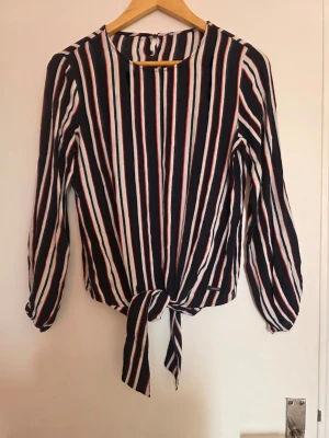 Randig blus med knytning framtill - Snygg blus med svarta, vita och röda vertikala ränder och knytning framtill. Blusen har rund halsringning och långa ärmar. Materialet känns lätt och luftigt, perfekt för en trendig look.