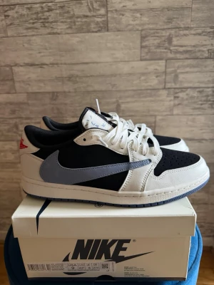 Nike Air Jordan 1 Low OG svart/vit - Nike Air Jordan 1 Low OG i svart och vitt med grå Swoosh och röda detaljer på hälen. Skon har en klassisk låg silhuett, perforerad tåbox och kommer med extra skosnören i blått och svart. Ovandelen är i läder och mocka för en stilren look.