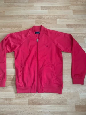 Ny träning jacka från Fred Perry - Snygg rosa/röd sweatshirts från Fred Perry med dragkedja framtill och två fickor. Klassisk ribbad mudd vid ärmslut och nederkant samt Fred Perry-logga broderad på bröstet. Perfekt för en avslappnad och sportig stil. Kom aldrig till användning.
