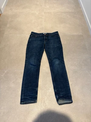 Mörkblå Jack&Jones jeans - Säljer ett par mörkblå jeans med slim/rak passform och klassisk femficksdesign. Jeansen har en enkel look med kontrastsömmar och knappgylf. Perfekta till vardags för en avslappnad stil.