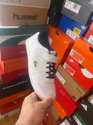 Vita Lacoste sneakers med kardborre - Fräscha vita sneakers från Lacoste med svart foder, svarta skosnören och en praktisk kardborreknäppning. Klassisk krokodil-logga på sidan och stilren design i syntetmaterial och textil. Perfekta för en clean och sportig look.