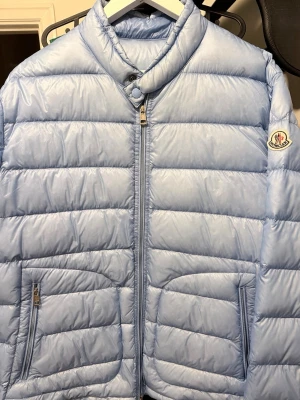 Ljusblå dunjacka från Moncler - Snygg babyblå dunjacka från Moncler med quiltad design och hög krage. Jackan har två dragkedjeförsedda fickor framtill och klassisk Moncler-logga på ärmen. Perfekt för  våren och riktigt stilren look. Storlek 5 dvs L-XL