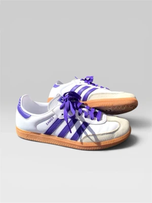 Adidas Samba vita/lila sneakers - Säljer ett par Adidas Samba sneakers i vitt och lila med klassiska tre ränder på sidan och gummisula. Skorna har lila detaljer, vita ovandelar i läder och mocka framtill. Perfekta för dig som gillar retrostil och sportig vibe.