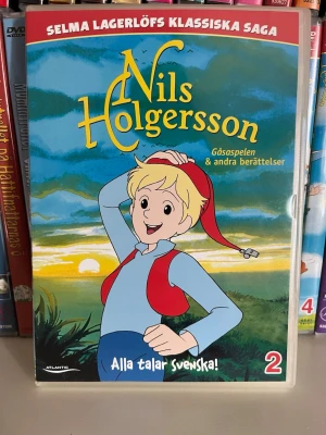 Nils Holgersson: Gåsspelet & andra berättelser - Nils Holgersson: Gåsspelet & andra berättelser är en svensk klassiker baserad på Selma Lagerlöfs saga. Omslaget är färgglatt med illustration av Nils i röd mössa och blå kläder, mot en landskapsbakgrund. Perfekt för dig som gillar äventyr och svensk litteratur.