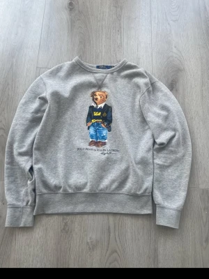 Grå sweatshirt Polo Bear Ralph Lauren - Snygg grå sweatshirt från Ralph Lauren med ikoniskt Polo Bear-motiv på bröstet. Tröjan har rund halsringning, långa ärmar och ribbade muddar. Perfekt för en avslappnad och trendig stil.