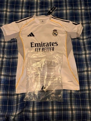 Real Madrid Mbappé matchtröja - Snygg vit Real Madrid matchtröja från Adidas med Mbappé och nummer 9 på ryggen. Tröjan har svarta detaljer, gula linjer längs sidorna och klubbmärke samt sponsortryck på bröstet. Perfekt för fotbollsfans som vill sticka ut på läktaren eller planen.