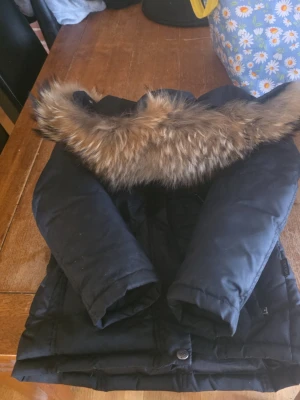 Svart dunjacka med fuskpäls - Snygg svart dunjacka med huva och avtagbar fuskpäls i bruna toner. Jackan har flera fickor framtill och dragkedja med knappslå. Perfekt för kalla vinterdagar och riktigt varm.
