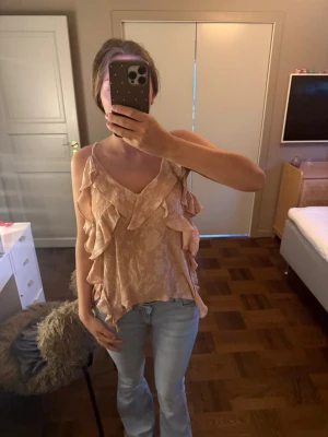 Beige volangblus med spetsdetaljer - Säljer en ljusbeige blus med volanger och tunna axelband. Blusen har v-ringning och är dekorerad med spetsmönster, vilket ger en romantisk och feminin känsla. Perfekt att styla med jeans eller kjol för en trendig look.