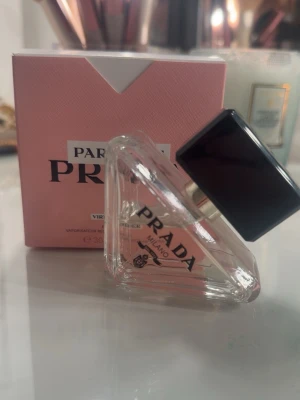 Prada Paradoxe Eau de Parfum 30ml - Prada Paradoxe Eau de Parfum i en unik, trekantig glasflaska med svart fyrkantigt lock. Flaskan har en elegant och modern design med tydlig Prada-logga. Förpackningen är ljusrosa med vita och svarta detaljer, vilket ger en lyxig känsla. Volymen är 30 ml.
