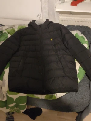 Svart pufferjacka från Lyle & Scott - Svart pufferjacka från Lyle & Scott med gul broderad logga på bröstet. Jackan har huva, dragkedja framtill och quiltad design som ger en klassisk pufferlook. Perfekt för kalla dagar och håller värmen bra tack vare det vadderade materialet.